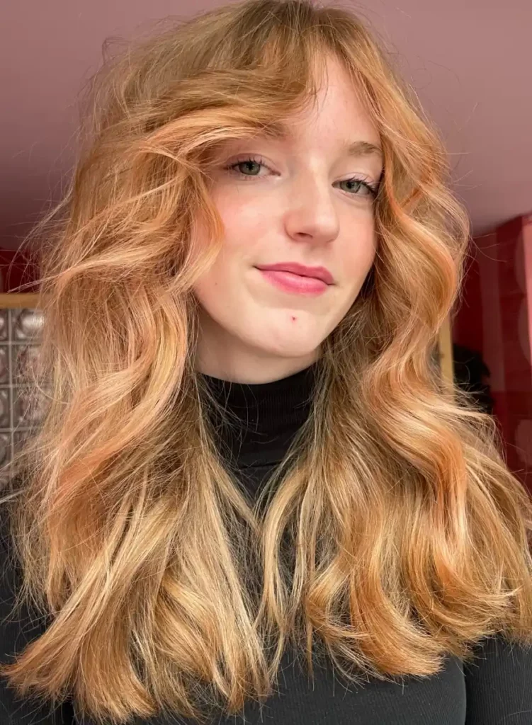 Vrouw met halflang, golvend haar in een warme blonde kleur met koperachtige toner, draagt een zwarte coltrui.
