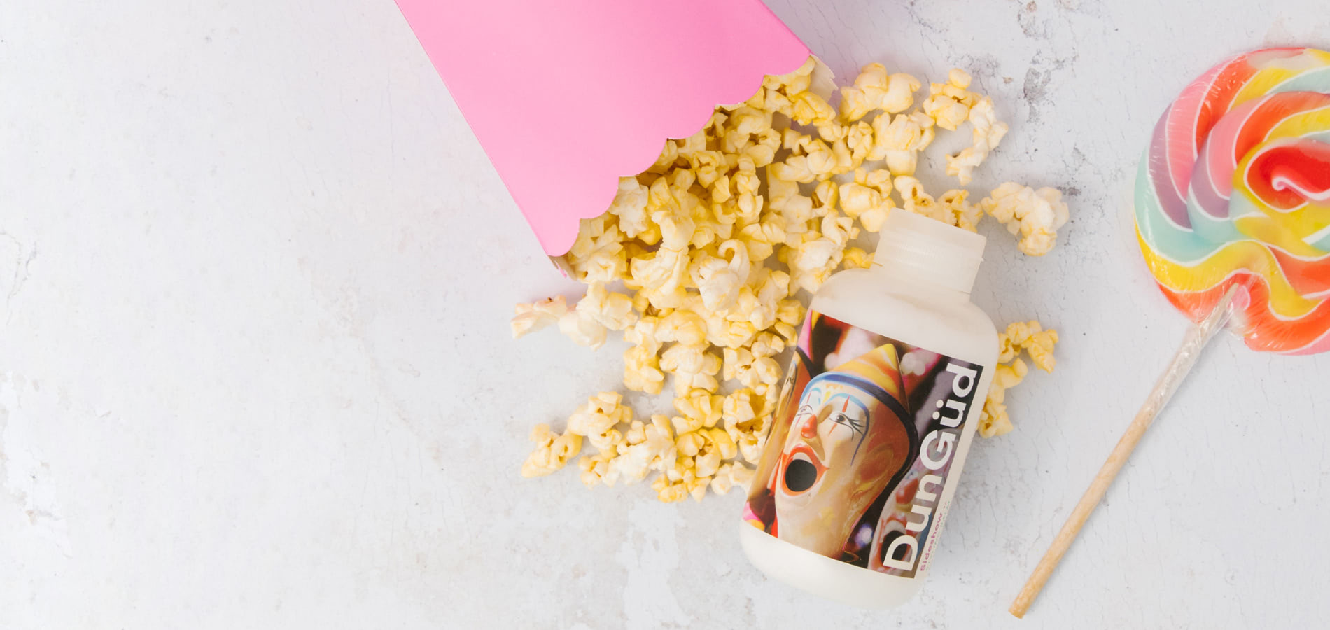 Popcorn, een kleurrijke lolly en een flesje met het label "DunGüd" met clownsafbeelding liggen op een lichte ondergrond.